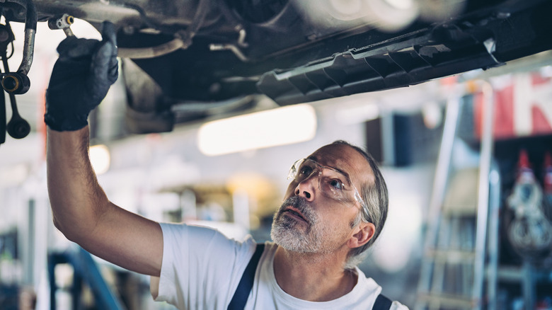 A man in safety glasses looks up at the underside of a car on a lift.