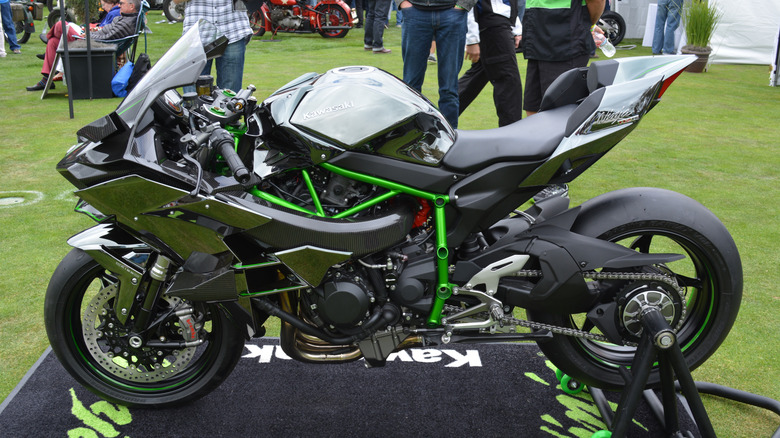 A darkly-colored Kawasaki Ninja H2 with green accents sits on a stand one a manicured lawn at an event.