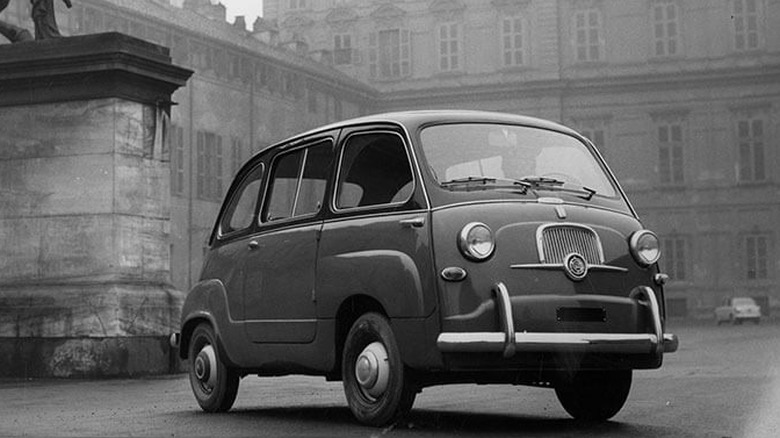 A black-and-white photograph of a vintage Fiat 600 Multipla parked in a misty European city square.