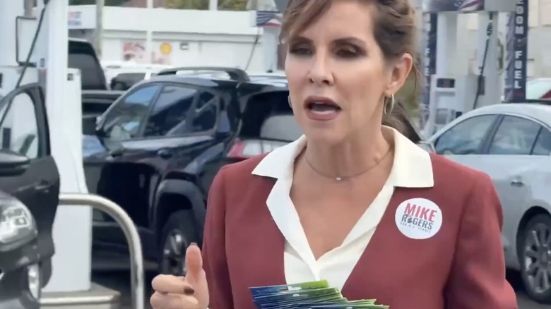 A woman in a red suit with a Mike Rogers sticker on her jacket holds a pile of gift cards in front of a gas station