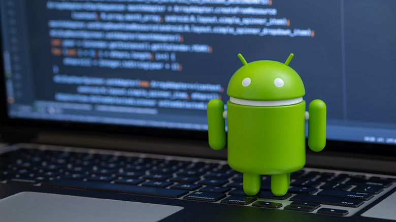 An Android toy standing on a computer keyboard with blurred code in the background