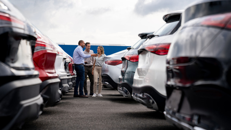 A small group discusses cars in a dealership parking lot.