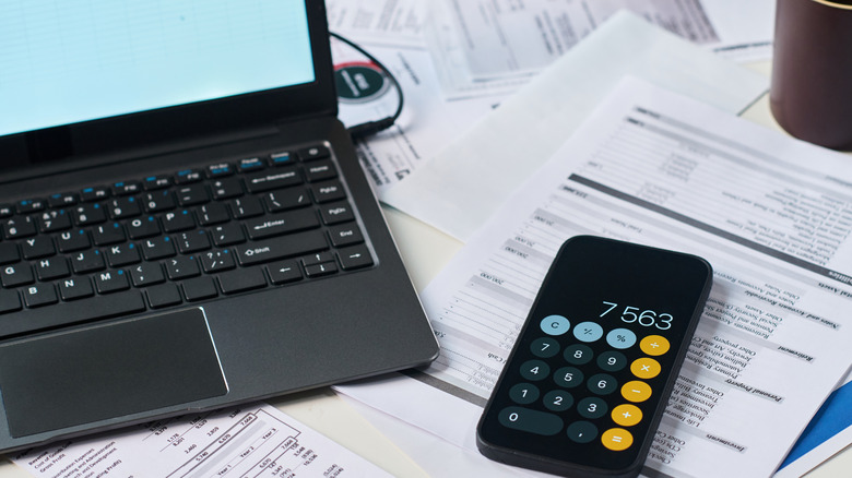 Papers, a laptop, and a phone with the calculator app open are spread on a desk.