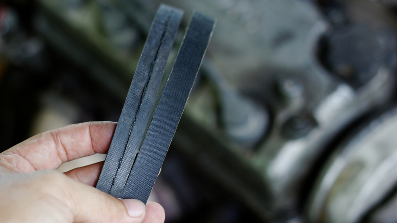 A hand holds a worn belt and a new one near an engine bay.
