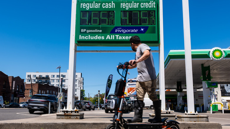 A man on an electric scooter looks at a gas price sign that reads "$4.65" for regular gas