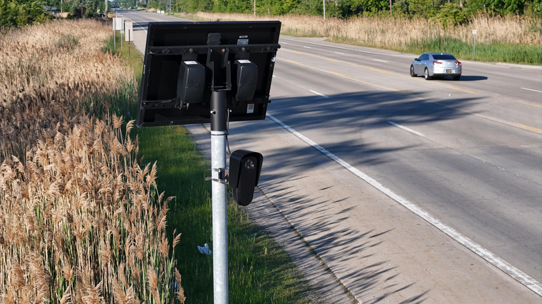 A Flock camera and solar panel mounted on top of a pole on the side of a four-lane highway