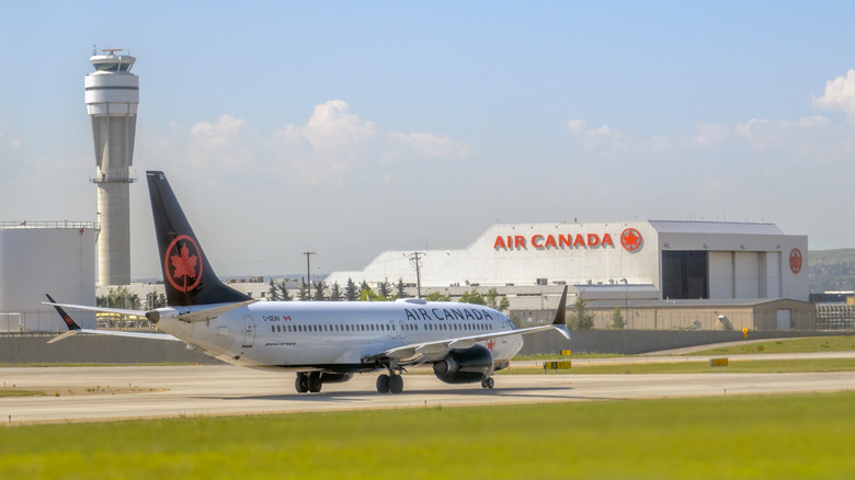 An Air Canada Boeing 737 MAX 8 aircraft taxis on the runway with an air traffic control tower and Air Canada hangar in the background.