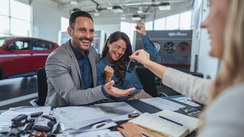 A smiling man and woman celebrate as a dealer hands them the keys during a positive car-buying experience. Papers and brochures spread across the desk in a modern showroom.