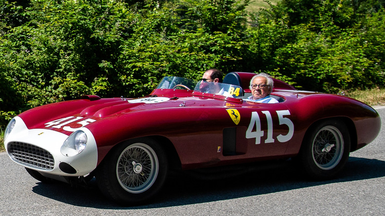 A red 1955 Ferrari 857 S with white numbers and grille area, and two men inside