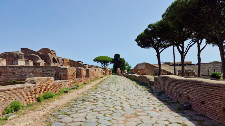 Another view of an ancient Roman cobblestone road going through Rome, Italy, with some older brick structures that are crumbling and trees along the path.