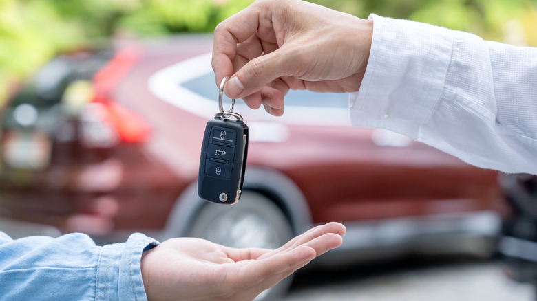 One person handing a car key to another, with a red car in the background.