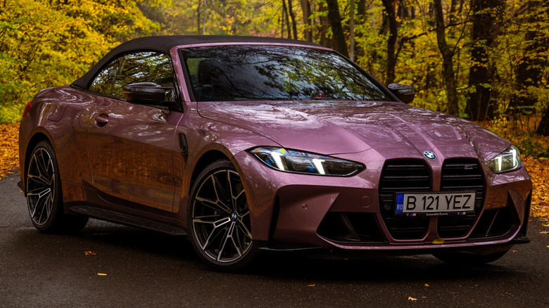 A violet 2024 BMW M4 convertible parked on a country road in an autumn setting.