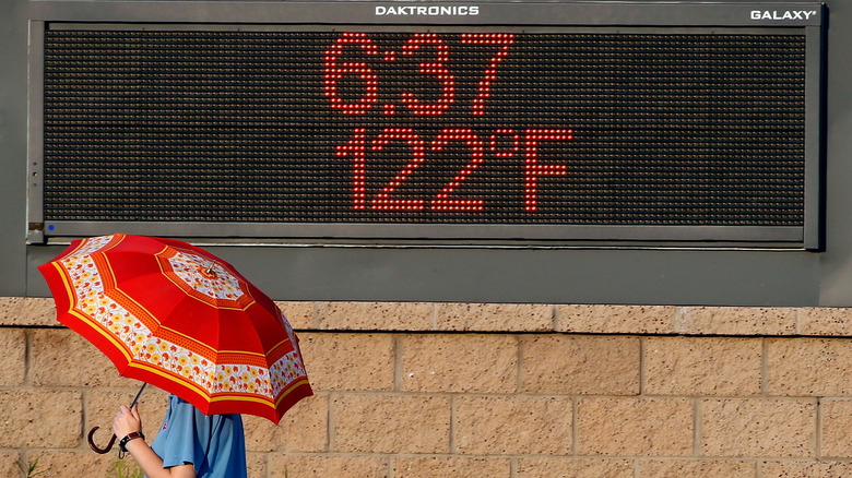 A man with an umbrella on a sunny day walking by a sign displaying a 122ºF temperature