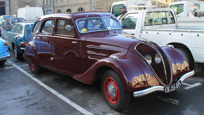 A wine-red Peugeot 402 sits parked in a busy lot with a brick building in the background