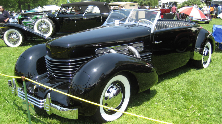 A black Cord 810 sits parked parked on grass at an auto show with other antique cars in the background.