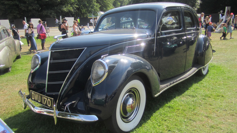 A black Lincoln-Zephyr sits parked on grass at a car show with many people in the background