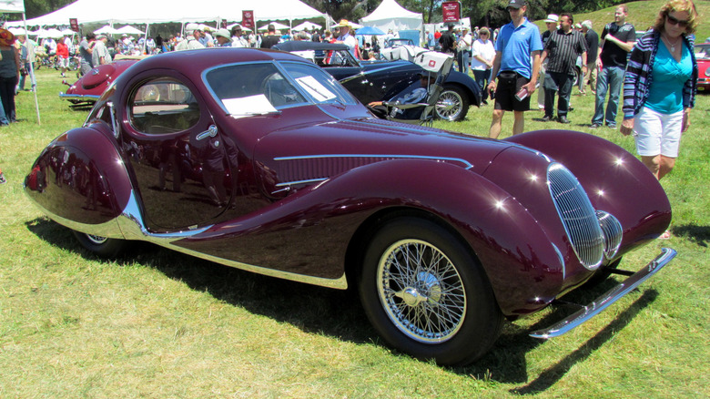 A purple Talbot-Lago T-150C-SS Teardrop Coupe sits parked on a concourse lawn as viewers admire it on a sunny day.