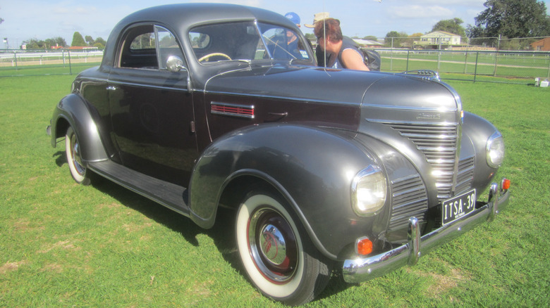 A two-toned Plymouth P8 Deluxe Coupe sits parked on the lawn in the sun as people inspect it closely in a small group.