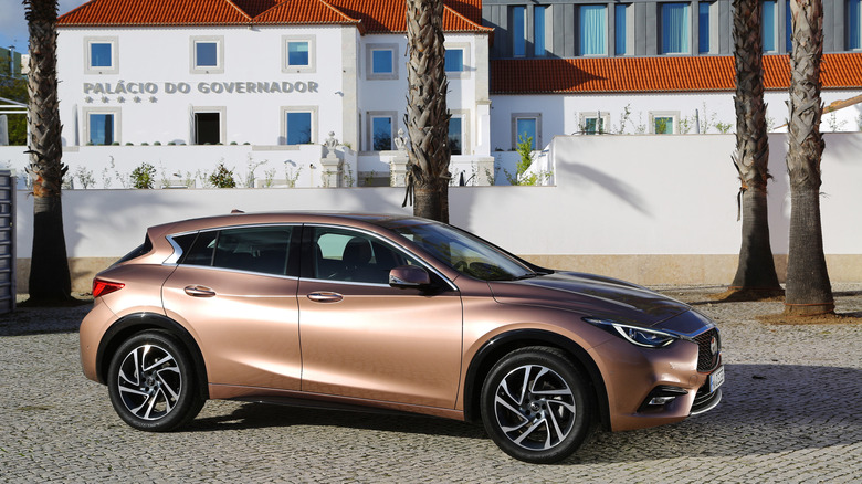 Side view of a copper Infiniti QX30 parked on bricks in front of palm trees and a white building