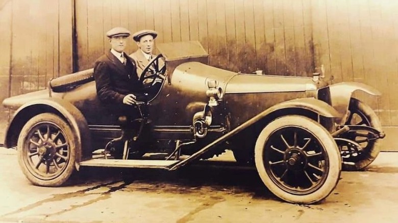 Two men in British caps and suits sitting in the Aston Martin Coal Scuttle car from a side view