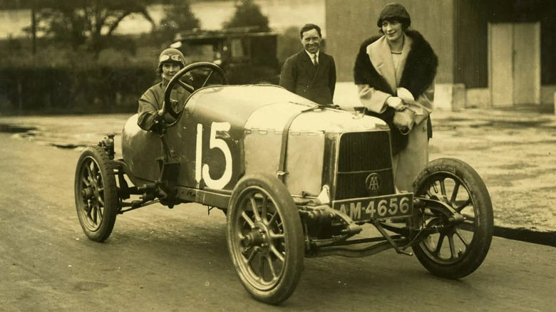 The Aston Martin Coal Scuttle car with "15" painted on the side, two well dressed people standing beside it, and a goggled driver behind the wheel