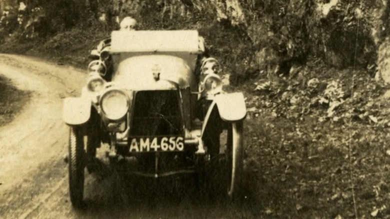 Aston Martin's first car, Coal Scuttle, seen from the front with one headlight driving toward the viewer on a dirt road next to a cliff face