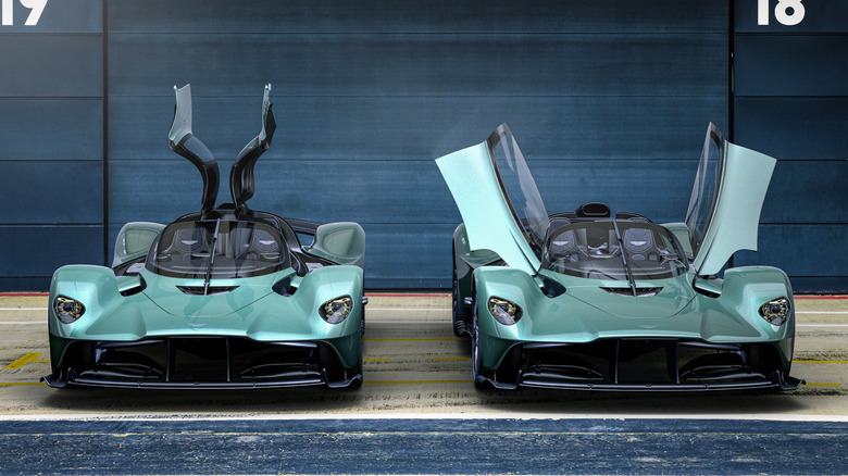 A head-on shot of an Aston Valkyrie coupe parked next to a Spyder both with their vertically opening doors up in front of a dark garage door