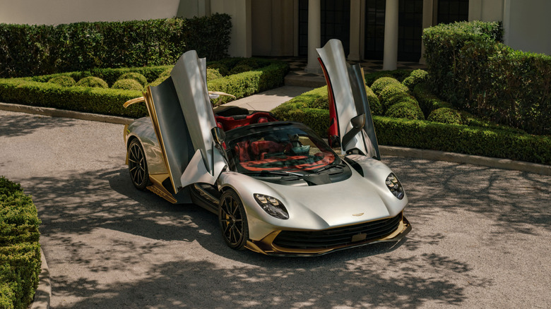A front three-quarters image of the silver-and-gold Valhalla Spider with its top off and doors up parked in front of a fancy house