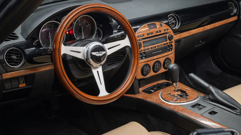 A close-up shot of the Miata's dashboard showing the wood-rimmed steering wheel with no airbag, but an Aston Martin emblem on the horn button