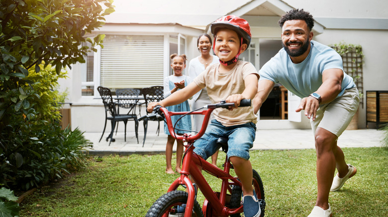 A proud dad teaches his young son to ride a bicycle  outside their home while wearing a helmet for safety.