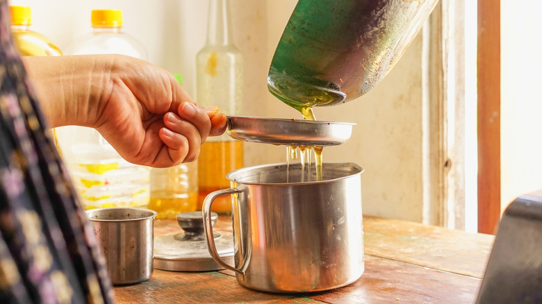 A person pouring used vegetable oil out of a pan, through a strainer, and collecting it in a pot