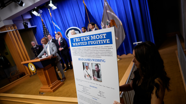 An aide displays a wanted poster during a news conference announcing the indictment of a former Olympic snowboarder Ryan Wedding, charging him with murder and money laundering in connection to a drug trafficking organization at the Justice Department on November 19, 2025 in Washington, DC.