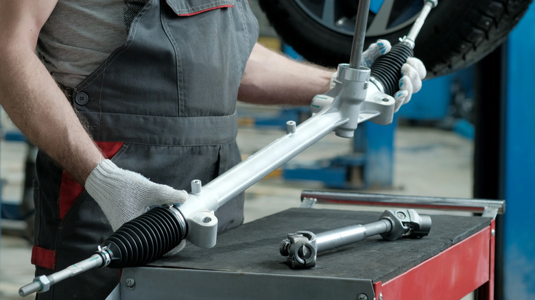 A mechanic holding a rack and pinion steering rack with gloved hands, tilting its left side.
