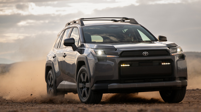 A front three quarters shot of a gray Toyota RAV4 Woodland edition driving through dirt with its headlights on