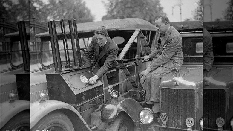 A man adjusts an old radio mounted to the side of an old car while another man sits on another car next to him