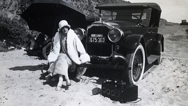 A woman sitting on the front bumper of a parked old car with a large old radio on the ground next to her