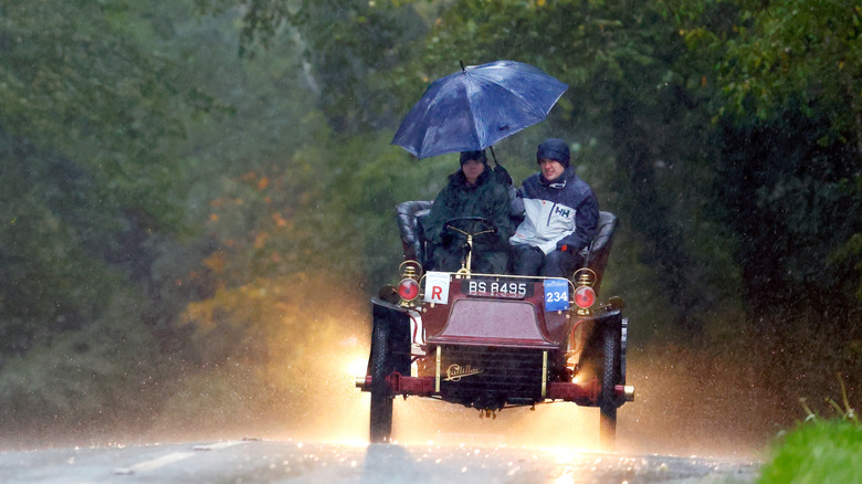 The driver and passenger of a 1904 Cadillac holding an umbrella while driving in the rain