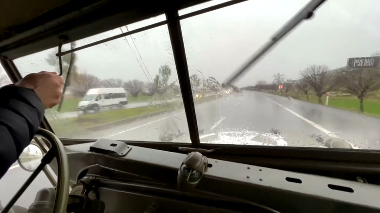A driver in a Willys Army Jeep operates the manual windshield wipers while driving in the rain on a highway.