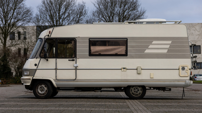 A vintage beige motorhome parked on a paved lot, framed by leafless winter trees and modern buildings in the background