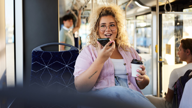 Larger blonde woman with glasses talks on her phone on speaker while riding a public bus.