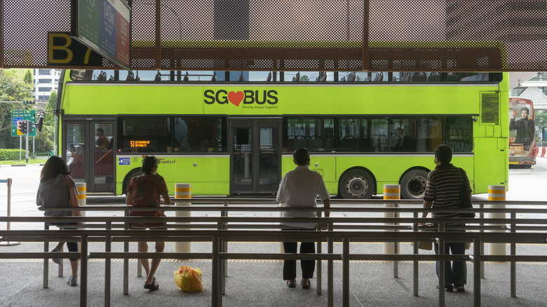 A group of commuters wait on a sidewalk with belted guides for a Signapore bus pulling up in the background of the city.