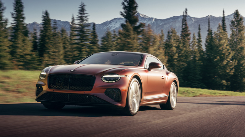 A front three-quarters shot of a 'Cricket Ball Satin' Continental GT driving on a rural road past trees and a mountainscape