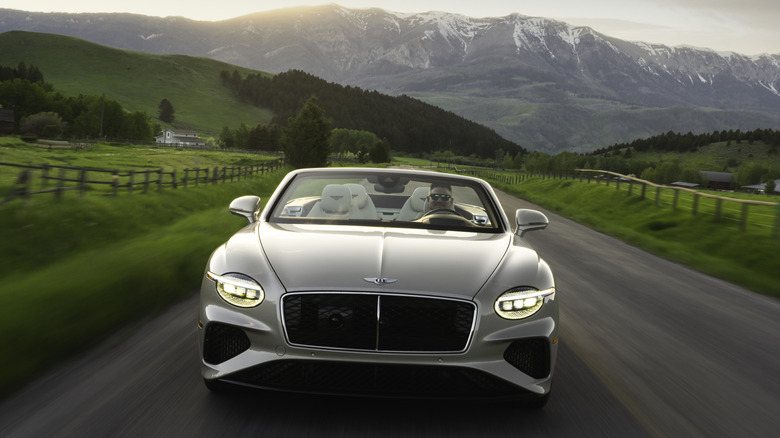 A head-on shot of a beige Continental GTC driving on a rural road surrounded by green grass in front of a mountain view at sunset