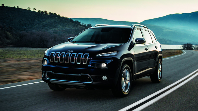 A Jeep Cherokee drives on a road through hills.