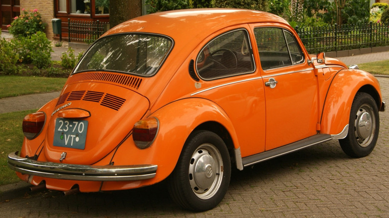 An orange 1973 Volkswagen Beetle parked on a brick street in front of a lawn, bushes, and brick storefront