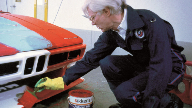 Andy Warhol brushes a line of red paint with a large paint brush across the front bumper of the fourth BMW Art Car.