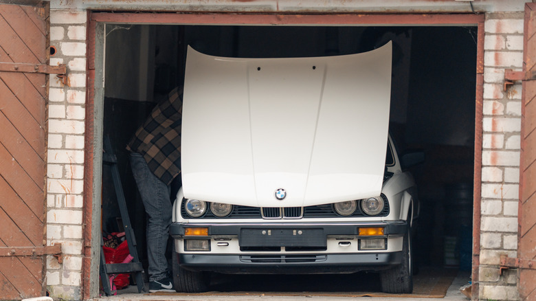 A man under the hood of white classic convertible BMW E30 car in the opened garage.