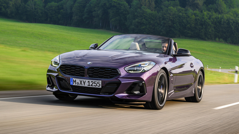A dark purple metallic BMW Z4 drives with the roof down on a country road, with green grass and trees in the background.