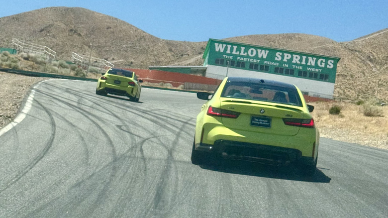 A photo out the windshield of a car on the track showing the back of a highlighter yellow M3 following a highlighter yellow M2 driving uphill toward a big green Willow Springs sign