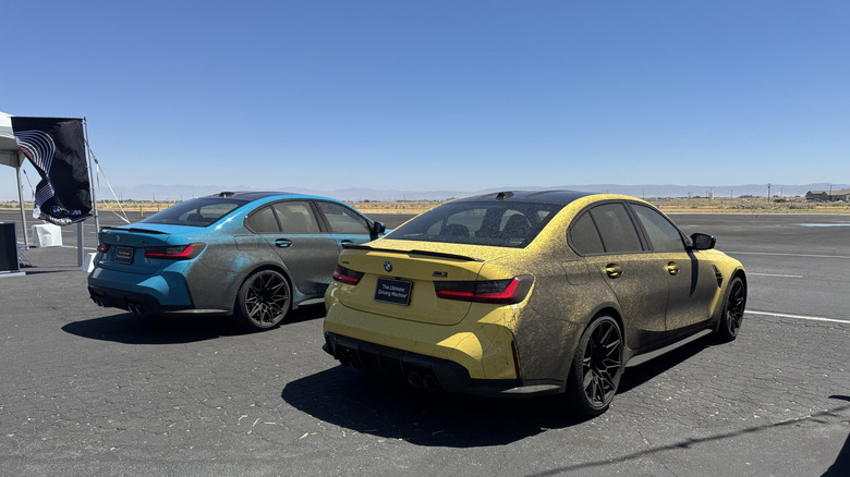 A photo of a light blue and light yellow M3 parked next to eachother on blacktop while the sides are completely covered in black overspray